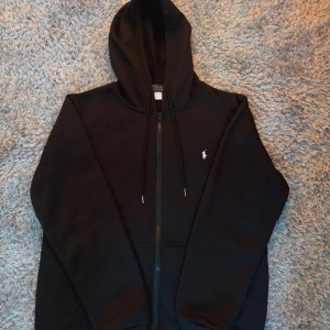 Svart hoodie från Polo Ralph Lauren - Svart hoodie från Polo Ralph Lauren med dragkedja och huva. Klassisk design med vit broderad logga på bröstet och snörning i huvan. Perfekt för en chill och stilren look. Priser går att diskuteras!