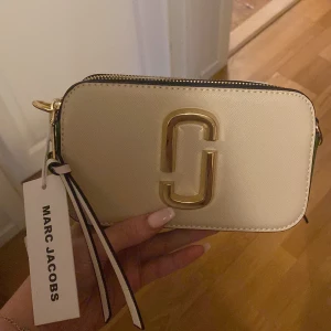 Vit axelväska från Marc Jacobs - Snygg och stilren axelväska från Marc Jacobs i vit färg med guldiga detaljer och dubbla dragkedjor. Väskan har en bred axelrem med loggan på. Perfekt storlek för det viktigaste och ger en trendig touch till din outfit. Jag har aldrig använt den eftersom att jag har så många väskor så därför har jag aldrig fått ett tillfälle för att använda den