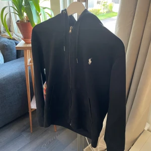 Svart hoodie från Polo Ralph Lauren - Svart hoodie från Polo Ralph Lauren med dragkedja och huva. Klassisk logga broderad på bröstet i vitt. Tröjan har långa ärmar, snörning i huvan och två fickor framtill. Perfekt för en chill och stilren look. Bara använd ett fåtal gånger, kan sänka priset vid snabb affär. Ny pris 2000kr 