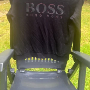 Svart Hugo Boss sweatshirt slim fit - Svart sweatshirt från Hugo Boss med stor BOSS-logga framtill. Slim fit-modell med rund hals och långa ärmar. Tröjan är gjord i mjuk bomull och har ribbade muddar vid hals, ärmslut och nederkant. Perfekt för dig som gillar stilrena och klassiska plagg.