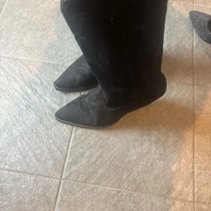 Svarta cowboy boots i mocka - Snygga svarta cowboy boots med spetsig tå och låg klack. Skorna är i mocka med klassiska broderade detaljer på skaftet. Perfekta för dig som vill ha en cool westernstil och matcha med jeans eller klänning.