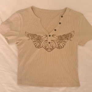 Beige topp med tryck och knappar - Snygg beige ribbad topp med kort ärm och coolt vingtryck framtill. Toppen har v-ringning med knappar som ger en extra detalj. Knappt använd! 