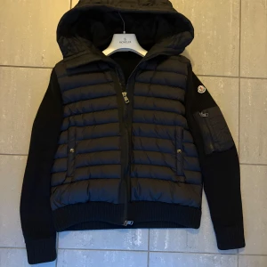 Svart cardigan från Moncler - Säljer en svart cardigan från Moncler med stickade ärmar och huva. Jackan har dragkedja framtill, fickor med knappar och en extra ficka med dragkedja på ärmen. Klassisk Moncler-logga på vänster ärm. Perfekt för kyliga dagar. Nfc och har haft denna jacka ca 1 och ett halvt år, supernöjd med den. 
