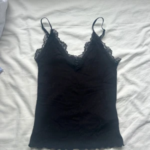Svart spetslinnetopp från Amisu XS - Svart linne från Amisu i storlek XS med smala axelband och v-ringning dekorerad med spets. Toppen är ribbad och har vågig kant nertill. Perfekt för att skapa en snygg lager-på-lager-look eller bära som den är.