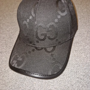 Svart keps från Gucci med GG-mönster - Snygg svart keps från Gucci med diskret GG-mönster över hela kepsen. Kepsen har böjd skärm och detaljer i svart skinn baktill. Tillverkad i ett tåligt tygmaterial med exklusiv känsla. Perfekt för dig som vill ha en stilren och lyxig accessoar.