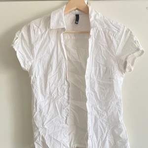 Vit kortärmad skjorta i bomull - Klassisk vit skjorta med korta ärmar och knappar framtill. Skjortan formar kroppen fint och är tillverkad i bomull, perfekt för en clean och enkel stil. Snygg krage och enkel design som funkar till många outfits.
