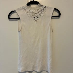Vit ärmlös topp med spetsdetalj - Supersöt vit ärmlös topp med ribbad struktur och vacker spets i blommigt mönster vid halsen och nacken. Perfekt för dig som gillar detaljer och vill ha något stilrent men ändå unikt. Snygg att styla med jeans eller kjol. Stoleken är xs men passar även s
