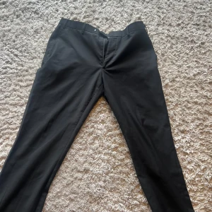 Svarta kostymbyxor från Dressmann - Svarta kostymbyxor från Dressmann i klassisk rak modell. Byxorna har hällor för bälte och är tillverkade i ett slätt tyg som känns dressat och stilrent. Perfekta för dig som vill ha en clean och enkel look. Så ser man på sista bilden att det är en fläck där som inte riktigt går bort den e på högra benet längst ner 