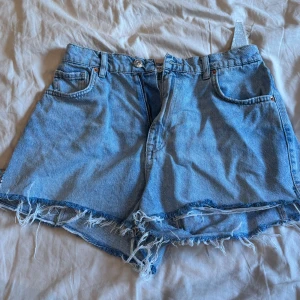 Blå jeansshorts från Pull&Bear - Snygga ljusblå jeansshorts från Pull&Bear med hög midja och fransig, rå kant. Cool design på fickorna med knapp och dragkedja framtill. Perfekta för sommaren och har en snygg passform som ger en chill vibe. Säljs då de är för små