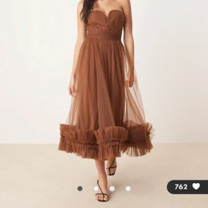 Brun axelbandslös tyllklänning - Superfin brun axelbandslös klänning i tyll med volangkant nertill. Klänningen har en markerad midja och ett romantiskt, flowy fall. Perfekt för dig som vill sticka ut med en elegant och trendig look. Klänningen har inte använts och har fortfarande prislappen på. 