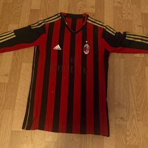 AC Milan långärmad Baresi tröja Adidas - Säljer en klassisk AC Milan långärmad fotbollströja från Adidas med Baresi och nummer 6 på ryggen. Tröjan har svarta och röda vertikala ränder, guldiga Adidas-ränder på axlarna och klubbmärke på bröstet. Perfekt för dig som älskar italiensk fotbollshistoria.