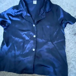 Mörkblå satinlik pjamas tröja från SHEIN - Snygg mörkblå pjamas tröja från SHEIN i satinliknande material. Tröjan har korta ärmar, klassisk krage, vita knappar framtill och en bröstficka. Perfekt för dig som gillar en lite glansig och stilren look.