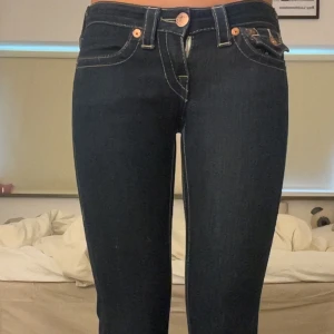 Mörkblå True Religion bootcut jeans - Säljer ett par mörkblå jeans från True Religion med bootcut passform och kontrasterande vita sömmar. Jeansen har låg midja, klassiska fickor med dekorativa lock och kopparfärgade knappar. Materialet är stretchigt denim som sitter snyggt och bekvämt. Gylfen är däremot sönder men går att laga, därav priset💞köpta second hand och säljs pga för korta.