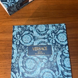 Versace Eros Eau de Toilette set - Lyxigt presentset från Versace med Eros Eau de Toilette och tillhörande body lotion. Förpackningen är blå med guldiga detaljer och har ett elegant, mönstrat blommotiv. Flaskan och tuben är båda i metallicblått med Versace-loggan. Perfekt för dig som gillar exklusiva dofter.