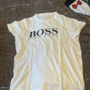 Vit Hugo Boss t-shirt med tryck - Vit t-shirt från Hugo Boss med stort svart BOSS-tryck på bröstet. Klassisk rund hals och korta ärmar. Mjuk och skön bomullskvalitet som känns chill och enkel att matcha med jeans eller shorts. Perfekt för dig som gillar stilrena logotoplagg.