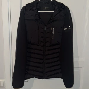 Svart Hybridjacja från Sail Racing - Nypris: 3500kr. Svart Hybrid Jacka från Sail Racing med huva och snygga quiltade paneler. Jackan har dragkedja framtill, två bröstfickor med dragkedja och logga på ärmen. Perfekt för kyliga dagar och har en sportig vibe. Materialet är polyester och syntet.