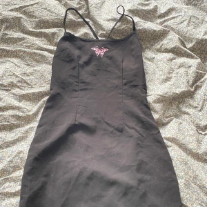 Svart kort klänning med öppen rygg. - Säljer en svart kort klänning med tunna axelband och snygg öppen rygg med snörning. Framtill finns ett litet rosa tryck med fjärilsmotiv. Klänningen har en slim passform och är perfekt för dig som gillar stilrena detaljer och en lite edgy look.