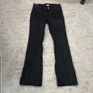 Svarta bootcut-byxor från Nelly - Snygga svarta byxor från Nelly med bootcut-ben och hög midja. Byxorna har dubbla knappar framtill och coola fickdetaljer på framsidan. Perfekta för dig som gillar en stilren look med lite edge.