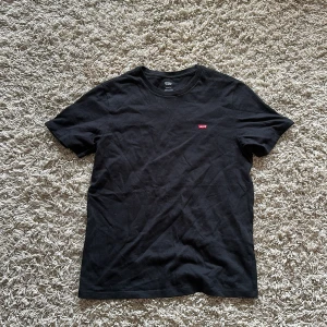 Svart Levi's t-shirt med logga - Svart t-shirt från Levi's med klassisk rund hals och korta ärmar. På bröstet sitter en liten röd Levi's-logga som ger en clean och stilren vibe. Materialet är mjuk bomull och passformen är normal, perfekt till jeans eller shorts.