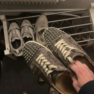 Christian Louboutin sneakers med nitar - Unika sneakers från Christian Louboutin i mörkgrått kamouflagemönster med coola silverfärgade nitar på tån. Skorna har klassisk röd sula, snörning och detaljer i skinn. Perfekta för dig som vill sticka ut med en edgy vibe.