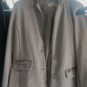 Grå kavaj med volangdetaljer - Snygg grå kavaj från Cream med två knappar framtill och dekorativa volangdetaljer på fickorna. Rak passform och klassisk krage, perfekt för en stilren look. Tidlös design som funkar till många outfits.