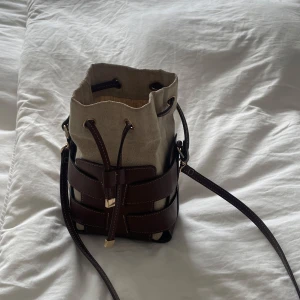 Beige och brun bucket bag från H&M - Säljer en snygg bucket bag från H&M i beige canvas med bruna skinnremmar och guldfärgade detaljer. Väskan har justerbar axelrem och stängs med snörning. Perfekt storlek för det viktigaste och ger en cool, avslappnad vibe.