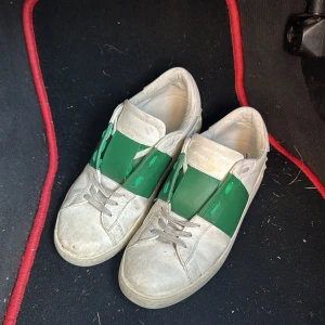 Valentino sneakers med grön detalj - Säljer ett par vita Valentino sneakers med bred grön rem över mitten och coola nitar baktill. Skorna har snörning och är tillverkade i skinn. Klassisk rund tå och platt sula. Perfekta för dig som vill sticka ut med en lyxig touch.