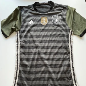 Tysklands randig fotbollströja Adidas - Snygg fotbollströja från Adidas med Tysklands landslagsmärke och FIFA 2014-emblem på bröstet. Tröjan är grå med svarta horisontella ränder och har olivgröna korta ärmar. Rund hals och vita detaljer längs sidorna. Perfekt för match eller träning. Köpt för 1500 kronor nu till ett pang pris för 299