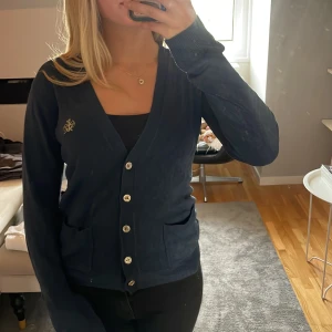 Mörkblå kofta från Gina Tricot - Mörkblå kofta från Gina Tricot med v-ringning, långa ärmar och snygga knappar framtill. Broderad gulddetalj på bröstet och två praktiska fickor. Perfekt för lager-på-lager och enkel att matcha med allt.
