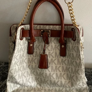 Michael Kors handväska - större modell  - Efterfrågad klassikern - snygg rymlig  damväska i vitt med konjakfägade- och guldiga detaljer. Väskan har MK-logga över hela ytan, två handtag och en axelrem med guldig kedja. Insidan har flera fack och har smart förvaring som rymmer bärbardator, böcker etc🌟. Perfekt för dig som gillar stilrena accessoarer. Hänglåset saknas - därav priset. Kan mötas i centrala- eller norra Stockholm för inbetalning och överlämning. 