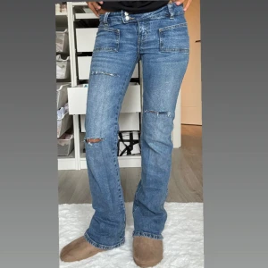 Blå bootcut jeans med slitningar - Snygga blå jeans med bootcut passform och slitna detaljer vid knäna. Jeansen har stora framfickor och klassisk femficksdesign. Perfekta för en avslappnad och trendig look.