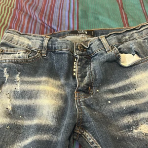 Blå distressed jeans från ICON XWAY - Säljer ett par blå jeans från ICON XWAY med slitna detaljer, blekta partier och färgstänk för en riktigt edgy look. Klassisk femficksmodell med orange sömmar och stor svartvit ICON-lapp bak. Perfekta för dig som gillar streetstyle.
