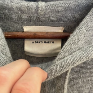 Grå hoodie i ull och kashmir A Day's March - Grå hoodie från A Day's March i storlek L. Tröjan är stickad i en lyxig blandning av 70% ull och 30% kashmir, har huva med snörning och långa ärmar. Perfekt för dig som gillar en stilren och avslappnad look.