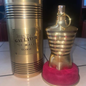 Jean Paul Gaultier Le Male Elixir - Exklusiv parfymflaska i guld från Jean Paul Gaultier, Le Male Elixir. Flaskan har en ikonisk kroppsliknande form med horisontella ränder och levereras med en matchande guldfärgad metallburk. Basen är klädd i röd sammet för en lyxig känsla.
