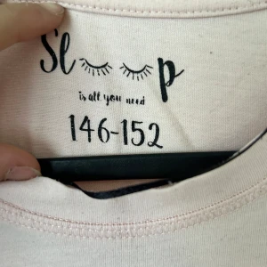 Ljusrosa t-shirtklänning med tryck - Säljer en ljusrosa t-shirtklänning från Sleep med trycket 'Sleep is all you need' och söta fransade ögon på bröstet. Klänningen har rund hals, korta ärmar och är gjord i mjuk bomull. Perfekt för chill dagar hemma eller som mysig nattklänning.