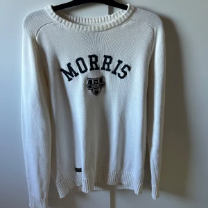 Vit stickad tröja från Morris Gents - Vit stickad tröja från Morris Gents med svart broderad logga och emblem på bröstet. Tröjan har rund halsringning, ribbade muddar och är långärmad. Perfekt för en clean och klassisk stil.