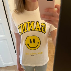 Vit Ganni t-shirt med gulligt tryck!!💛 - Vit t-shirt från Ganni med ett  gult smileytryck T-shirten har en klassisk rund hals och korta ärmar. Den  är i mjuk bomull och har en normal passform. Andbänd fåtal gånger så i mycket bra skick.  Perfekt statement-plagg med cool vibe!!🫰🏻⭐️