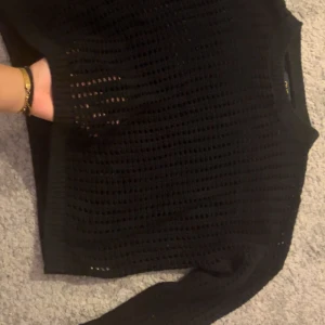 Svart stickad meshtröja - Säljer en svart stickad tröja med mesh-look och lång ärm. Tröjan har ett coolt hålmönster över hela och ribbade muddar vid ärmslut och nederkant. Perfekt att styla med ett linne under för en trendig vibe. Storlek M passar bra till S med