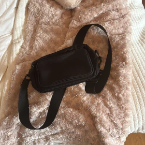 Svart axelväska med bred rem - En stilren svart axelväska med bred justerbar rem och praktiskt ytterfack med dragkedja. Perfekt för att bära det viktigaste och passar till många olika outfits. Enkel och clean design som funkar till allt.