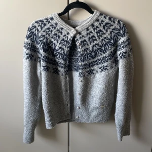 Grå kofta från Holly & Whyte - Säljer en mysig grå kofta från Holly & Whyte med ett vackert mönster i mörkblått runt oket. Koftan har långa ärmar och knäpps med knappar framtill. Perfekt för kyliga dagar!