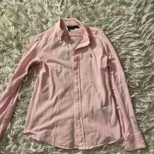 Rosa skjorta från Ralph Lauren - Snygg rosa skjorta från Ralph Lauren med klassisk krage och broderad logga på bröstet. Skjortan har långa ärmar och knappar framtill. Perfekt för en stilren look.