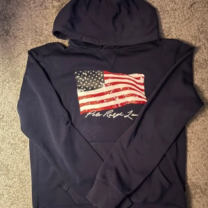 Mörkblå hoodie från Polo Ralph Lauren - Säljer en snygg mörkblå hoodie från Polo Ralph Lauren med en stor amerikansk flagga på bröstet. Tröjan har en klassisk känguruficka och en bekväm huva. Perfekt för en avslappnad stil.