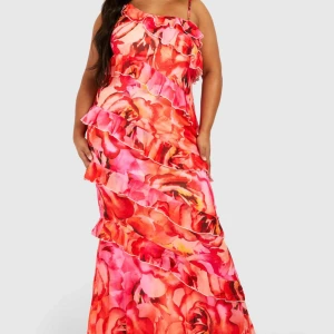 Färgglad långklänning med volanger - Säljer en färgglad långklänning med ett blommigt mönster i rosa och orange toner. Klänningen har asymmetriska axelband och volanger som ger en livfull look.