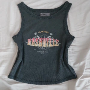 Ribbat linne Primark - Snygg grå ribbad linnetopp från Primark med tryck 'Nashville Glam Rock Unplugged Tour 1973' i färgerna röd, gul och vit. Perfekt för en avslappnad stil med en touch av rock.