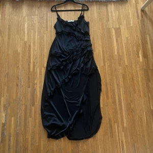 Svart långklänning från NA-KD - Elegant svart långklänning från NA-KD med volangdetaljer. Klänningen har smala axelband och en asymmetrisk design som ger en stilfull look. Perfekt för speciella tillfällen.