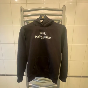 Svart hoodie från Peak Performance - Säljer en stilren svart hoodie från Peak Performance med vit logotyp på bröstet. Tröjan har en klassisk design med huva och långa ärmar. Perfekt för en avslappnad look.