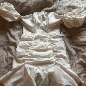 Vit studentklänning Nelly  - Säljer min backup studentklänning som aldrig kom till användning klänning från Love Triangle med puffiga ärmar och smockdetaljer på ryggen. Klänningen har en figurnära passform med volangdetaljer vid fållen. Perfekt för speciella tillfällen!