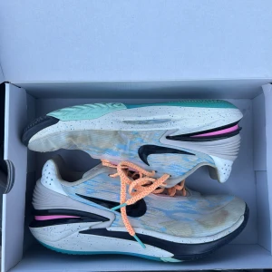 Basketskor - Säljer ett par Nike GT Cut 2 med en unik design. Skorna har en vit bas med svarta och turkosa detaljer samt orange snörning. Snörena är bytta därav färgen. Sulan är mönstrad med turkos och rosa inslag. Perfekta för träning eller vardagsbruk.