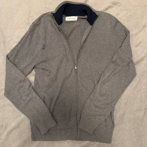 Grå kofta med dragkedja från Fire One - Snygg och stilren grå kofta från pire One med dragkedja och ribbade muddar. Perfekt för lager-på-lager under kyligare dagar. Den har en klassisk design med en mörkare krage som ger en fin kontrast.