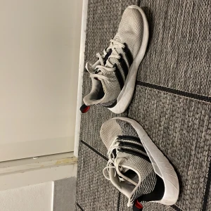 Vita och svarta sneakers från Adidas - Säljer ett par snygga vita sneakers från Adidas med svarta detaljer och klassiska ränder. Skorna har snörning och en bekväm sula, perfekt för vardagsbruk. De är tillverkade i syntetmaterial och har en modern design.